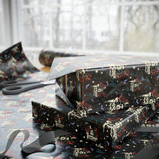 Gift boxes wrapped in Svengoolie holiday wreath wrapping paper stacked on a table beside black scissors and black ribbon.