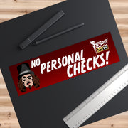 Overhead view of the red Svengoolie bumper sticker with 'NO PERSONAL CHECKS!' on a black surface beside a pen and ruler, showing its size.