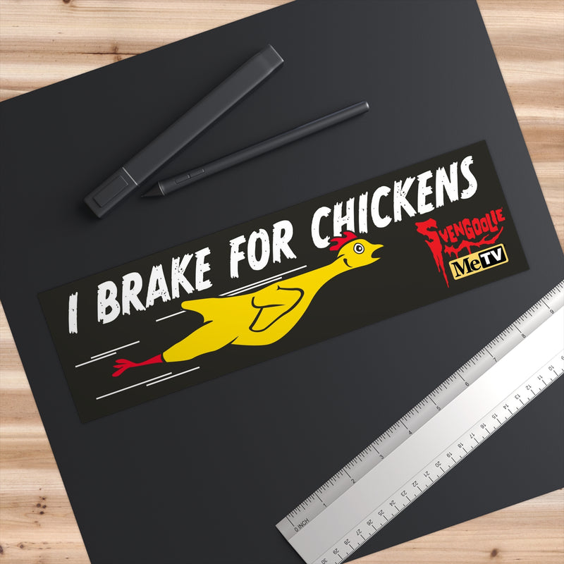 Overhead view of the I Brake for Chickens bumper sticker on a black mat beside a pen and ruler, showing its size.