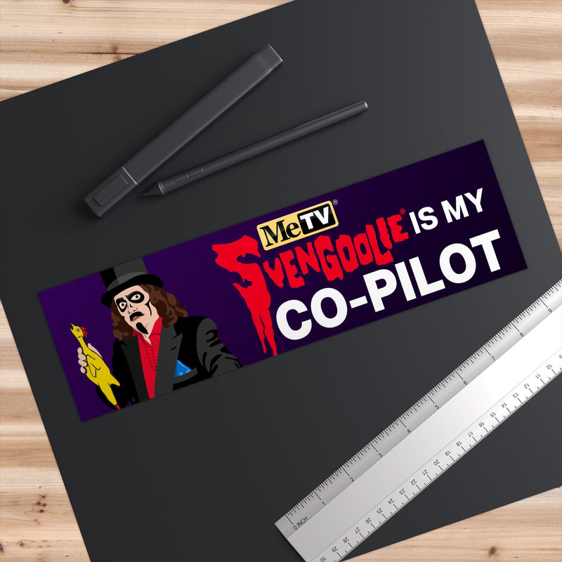 Svengoolie Is My Co-Pilot bumper sticker on a dark desk beside a pen and ruler, showing its size.