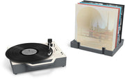 Angled view of gray and cream Victrola Re-Spin record player with a black vinyl record on the platter, beside a gray rack holding albums