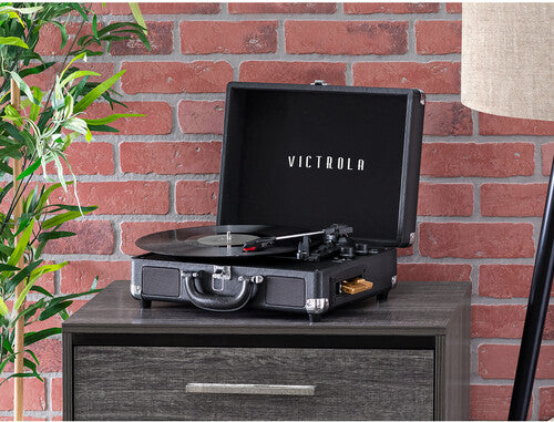 Black Victrola record player with its lid open and a record playing, set on a gray nightstand in front of a red brick wall.