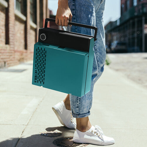 Model carrying the blue Victrola Revolution GO by its black handle on a city sidewalk, with the vertical Victrola logo visible on the side.