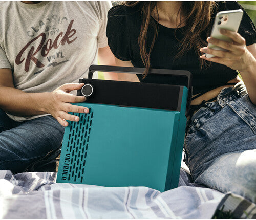 Two people seated outdoors with the closed blue record player on their laps, with one person holding its black carry handle.