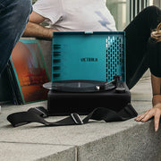 Lifestyle shot of the open blue record player with its black shoulder strap on a concrete step, with two people seated beside it.