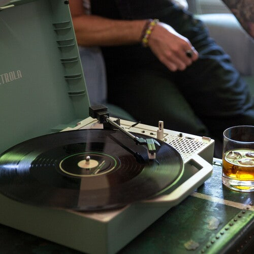 Angled view of basil green Victrola record player with the lid open and a vinyl record on the platter, beside a glass of amber drink.