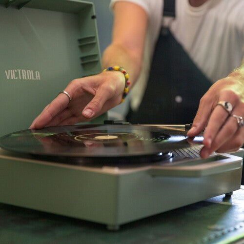 Close-up of hands placing a vinyl record on a basil green Victrola turntable, with the text 'VICTROLA' in light lettering on the lid.