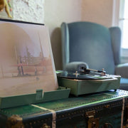 Basil green record player on a vintage trunk in a living room, with an album leaning in a stand beside it and a blue armchair behind.
