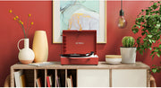 Red record player with lid open on a wooden sideboard in a styled room, beside a vase, plant, books, and bowl.