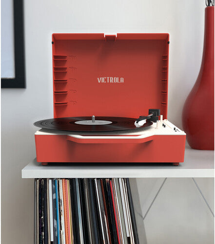Red record player with lid open and 'VICTROLA' printed on the lid, sitting on a shelf above a stack of vinyl records.