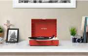 Red suitcase record player with lid open on a white shelf, with 'VICTROLA' printed on the lid, beside a framed photo and camera.