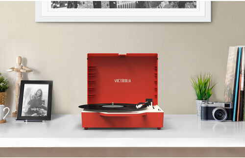 Red suitcase record player with lid open on a white shelf, with 'VICTROLA' printed on the lid, beside a framed photo and camera.