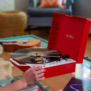 Hand with pink nail polish placing a record on the open red record player set on a glass coffee table in a living room.