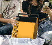 Two people sitting outdoors holding the closed citrus yellow Victrola Revolution GO portable record player with its carry handle, showing the vertical grille on its side.