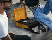 Person sitting on concrete steps beside the open orange Victrola Revolution GO record player with a record playing and its shoulder strap resting on the steps.