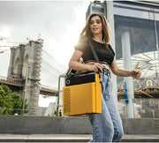 Model carrying the orange and black Victrola Revolution GO record player by its handle on a city street near a bridge.