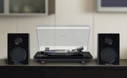 Studebaker Studio 88 turntable with the clear dust cover raised, shown between two black speakers on a wooden console in a living room.