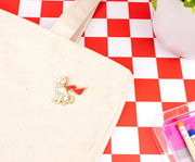 Krypto enamel pin on a cream canvas tote bag beside a red and white checkered tabletop with a plant.