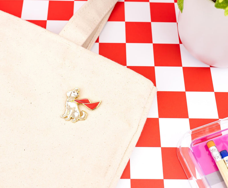 Krypto enamel pin on a cream canvas tote bag beside a red and white checkered tabletop with a plant.