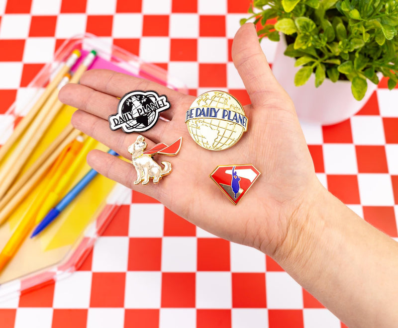 Hand holding Krypto enamel pin with three Daily Planet pins, including black 'DAILY PLANET' and gold 'THE DAILY PLANET' globes and a Superman shield.