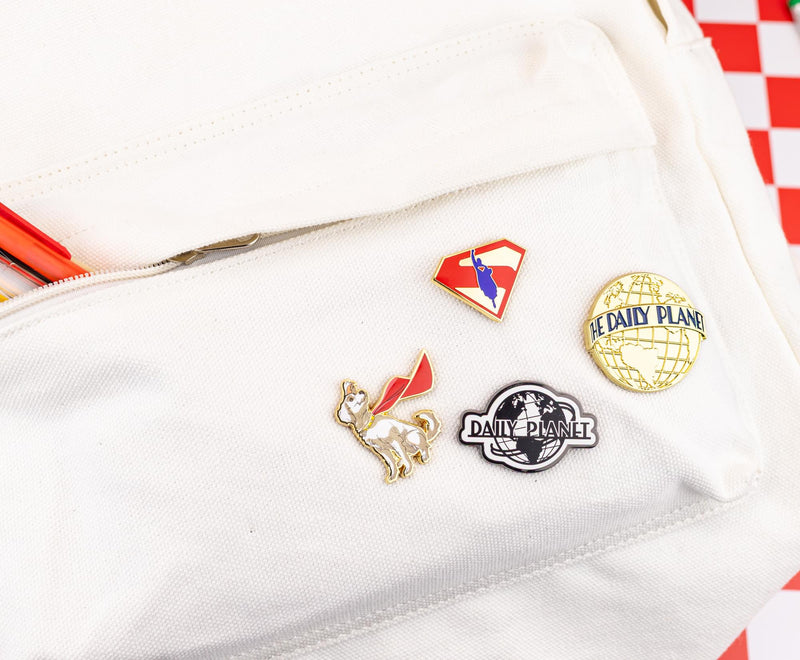 Krypto enamel pin on a cream backpack with Superman shield and Daily Planet pins nearby.