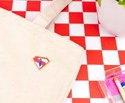 Superman flying logo pin attached to a cream canvas tote bag on a red and white checkered surface beside a potted plant.