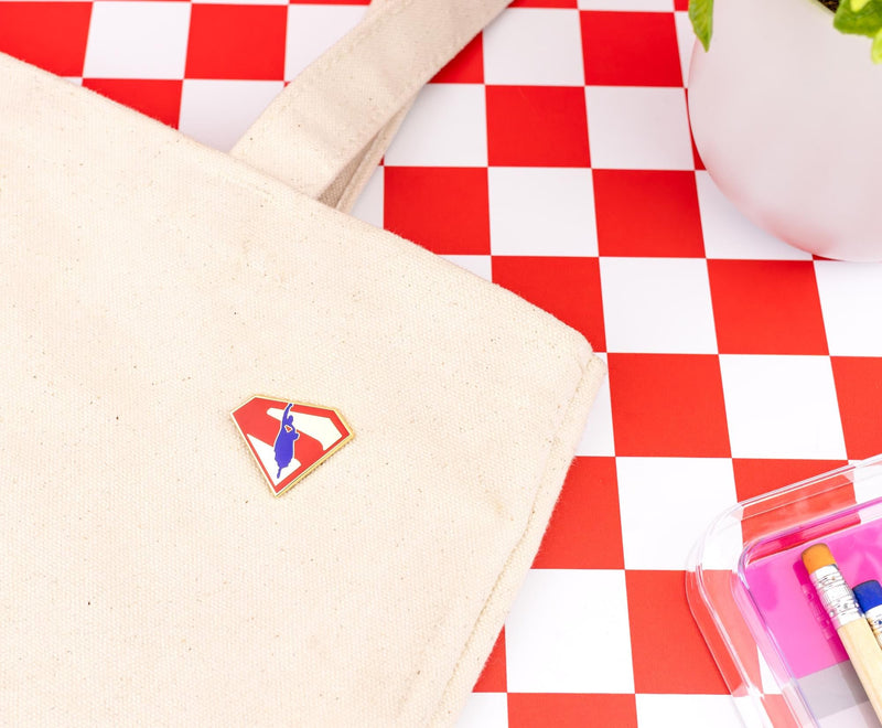 Superman flying logo pin attached to a cream canvas tote bag on a red and white checkered surface beside a potted plant.