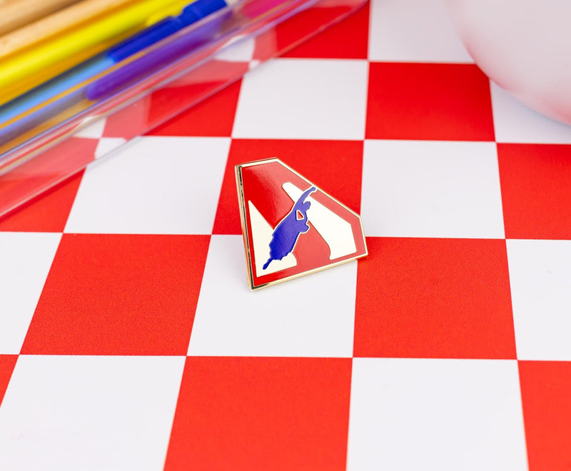 Angled close-up of the Superman flying logo pin on a red and white checkered surface, with markers in the background.
