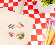 Four pins arranged on a cream tote bag, with the Superman flying logo pin at lower left beside a The Daily Planet globe pin.