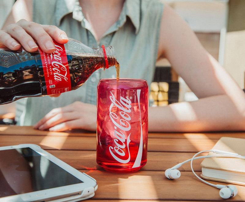 Coca-Cola being poured from a bottle into the red Coca-Cola glass cup on a wooden table, showing the white script logo on the cup.