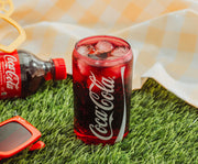 Red Coca-Cola glass cup filled with ice and soda on grass beside a bottle, showing the white Coca-Cola script logo along its side.
