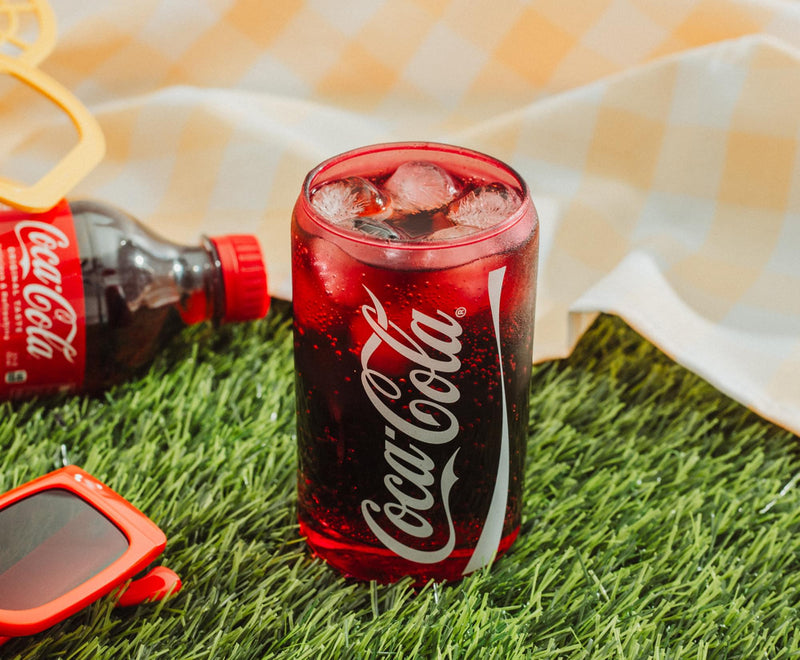 Red Coca-Cola glass cup filled with ice and soda on grass beside a bottle, showing the white Coca-Cola script logo along its side.