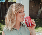 Model wearing a sleeveless denim shirt and holding the red Coca-Cola glass cup to her lips, with the white logo facing outward.