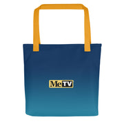 Navy-to-teal gradient tote bag with gold handles featuring the MeTV logo in black and gold centered on the front, with the text 'MeTV' in black and white on a gold label.