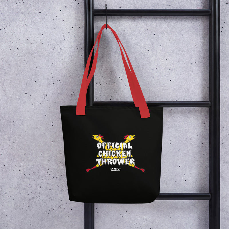 Black tote bag with red handles hanging on a black ladder, featuring two crossed yellow chickens with red combs and the text 'Official Chicken Thrower' in white above a small MeTV logo.