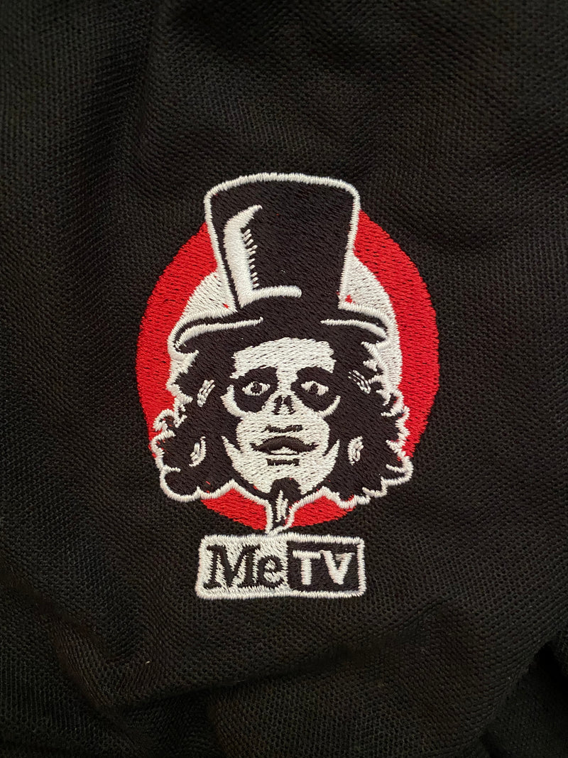 Close up view of Black polo shirt with small embroidered Svengoolie logo on chest: "MeTV" logo below.
