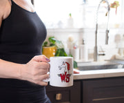 White ceramic mug with a red and black horse head above a large red 'WS' logo with black wings, held by a woman in a kitchen.