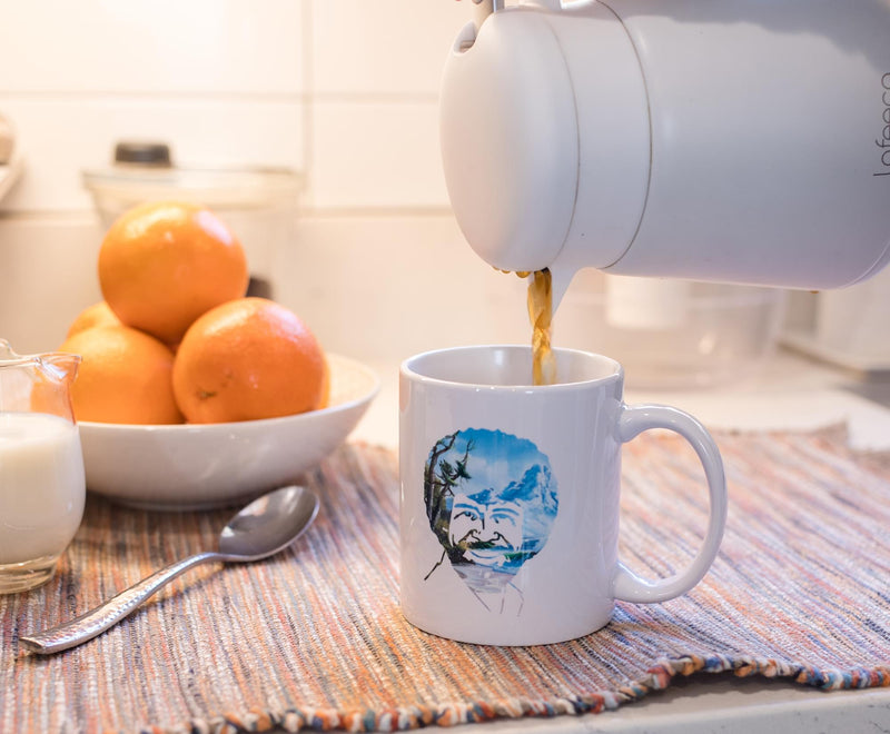 White ceramic mug with a circular printed portrait of Bob Ross, smiling, with a blue sky and mountain landscape behind him, being filled with coffee.