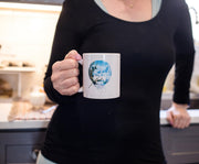 White ceramic mug held by a woman in a kitchen, printed with a blue-toned illustration of Bob Ross's face framed by clouds and evergreen trees.