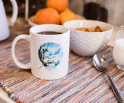 White ceramic mug of coffee with a circular print of Bob Ross's face blended into a blue mountain landscape with pine trees