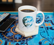 White ceramic mug with a circular blue-toned print of Bob Ross's face framed by pine trees and snowy mountains, holding black coffee.