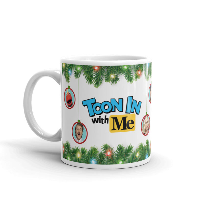 White ceramic mug with green pine garland with string lights, featuring round ornaments with faces and the text "TOON IN with Me" in blue and yellow.